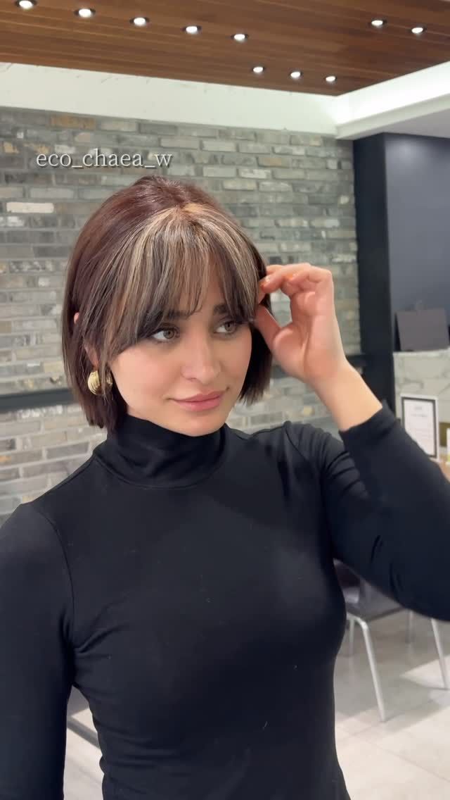 Woman in a black turtleneck adjusting her hair in a modern indoor lobby with stone walls.