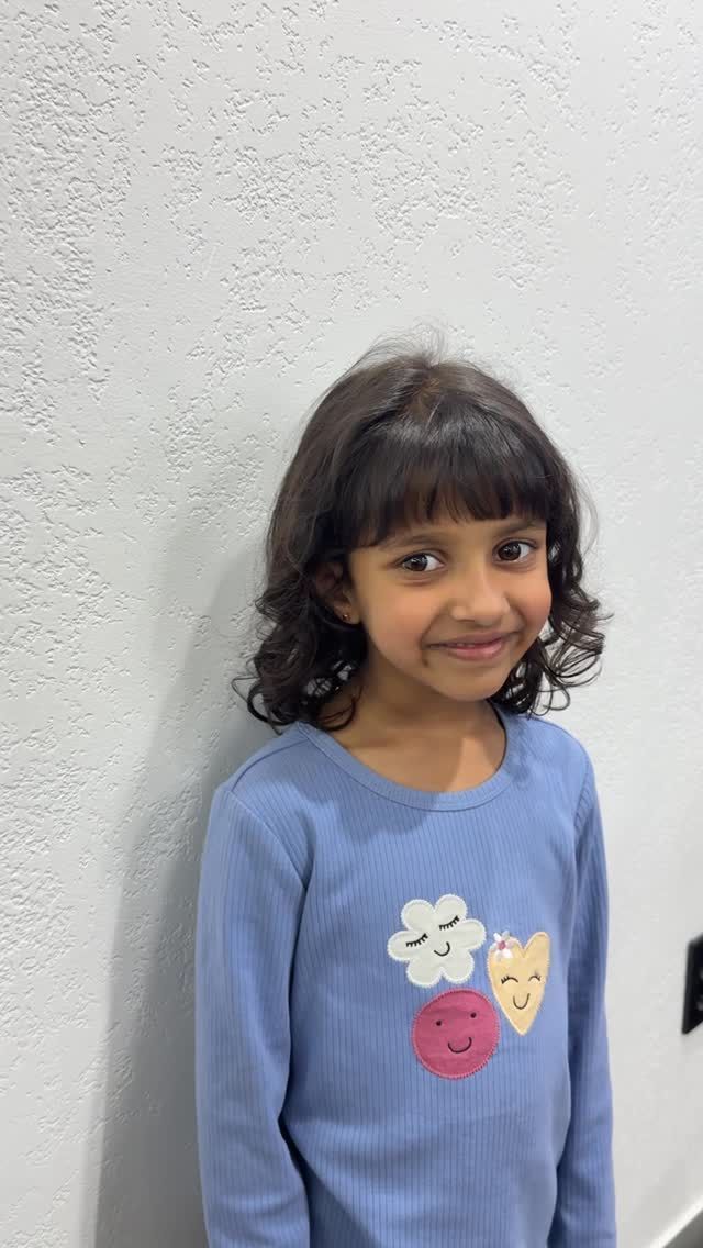 Young child standing against a white wall, smiling in a blue shirt with colorful heart designs.