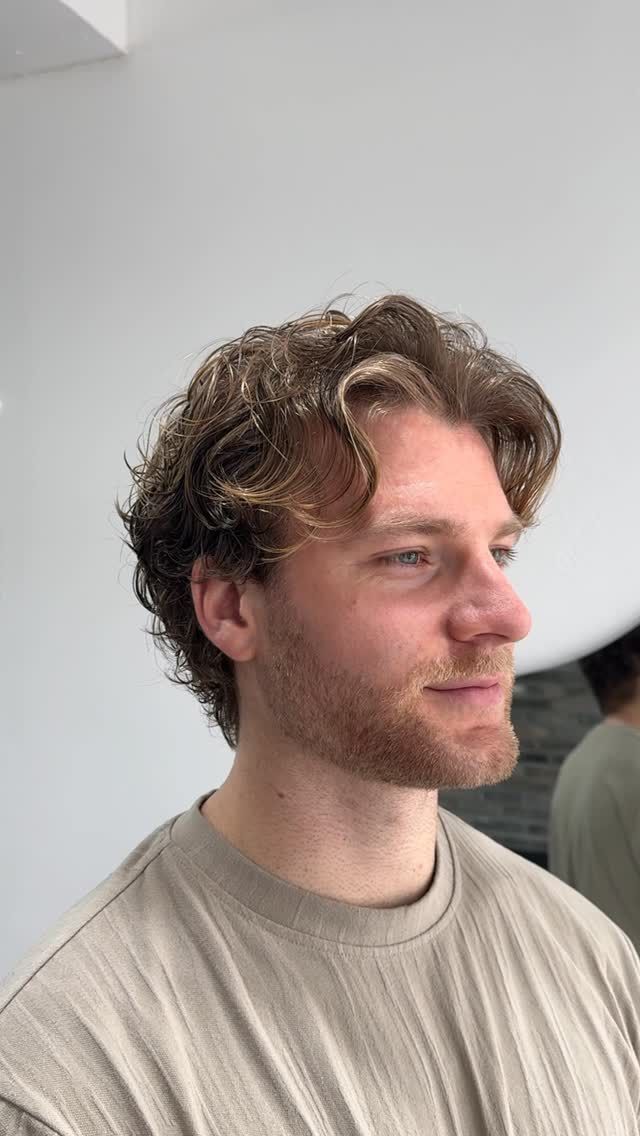 Side-profile portrait of a man with curly hair and a beige shirt against a plain white wall.