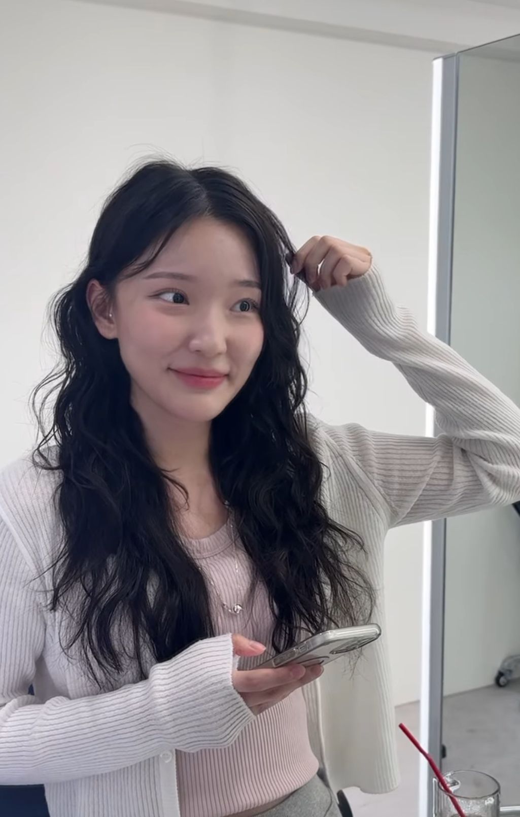 Woman in a white sweater adjusting her hair, holding a phone and smiling in front of a mirror.