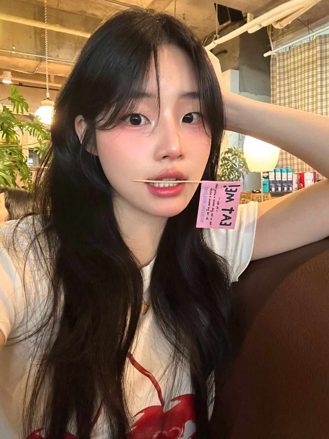 Young woman with long dark hair holding a pink tag, seated indoors in a café, gazing at the camera.