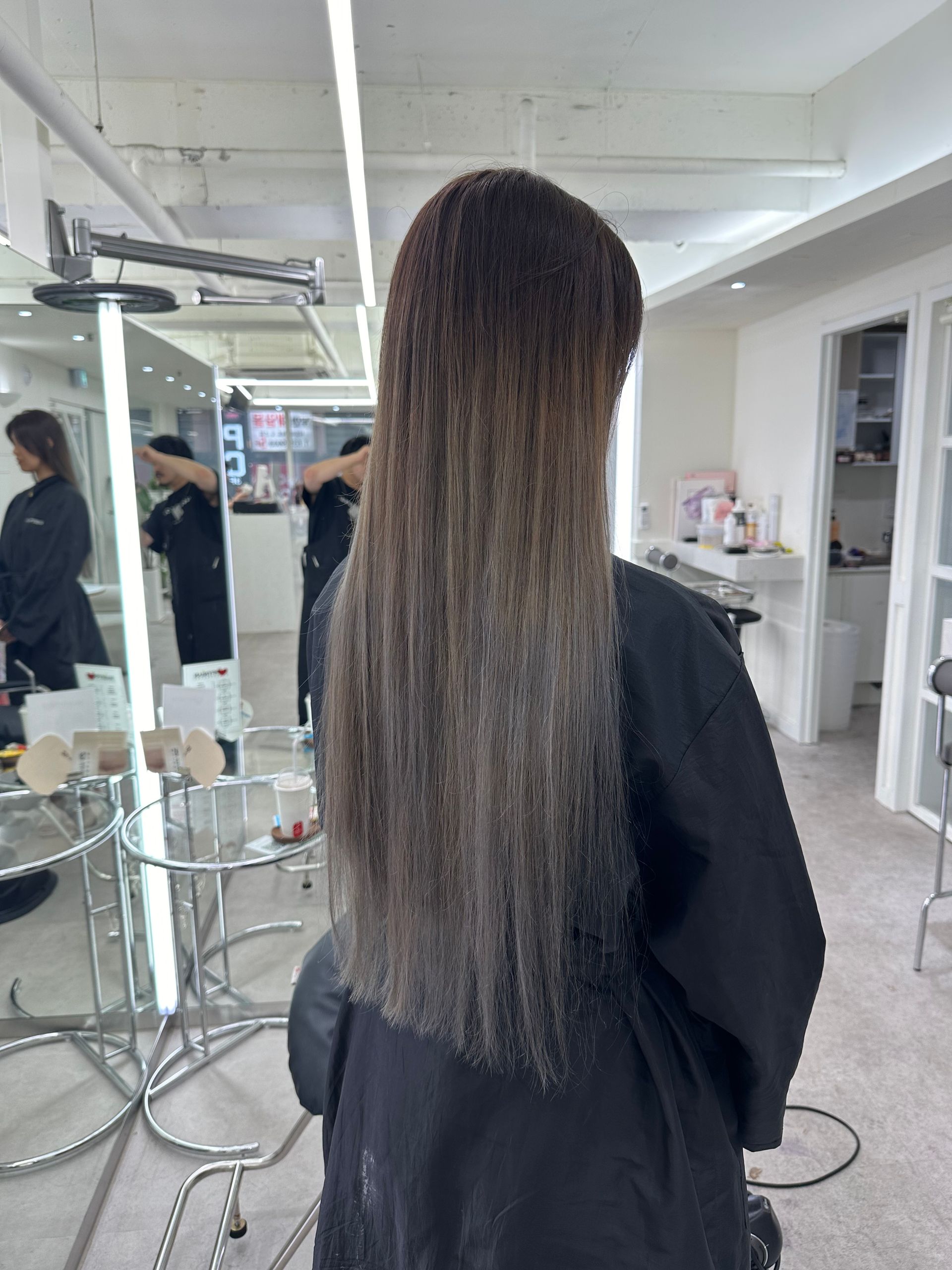 Back view of a person with long straight hair in a salon mirror area, wearing a black cape