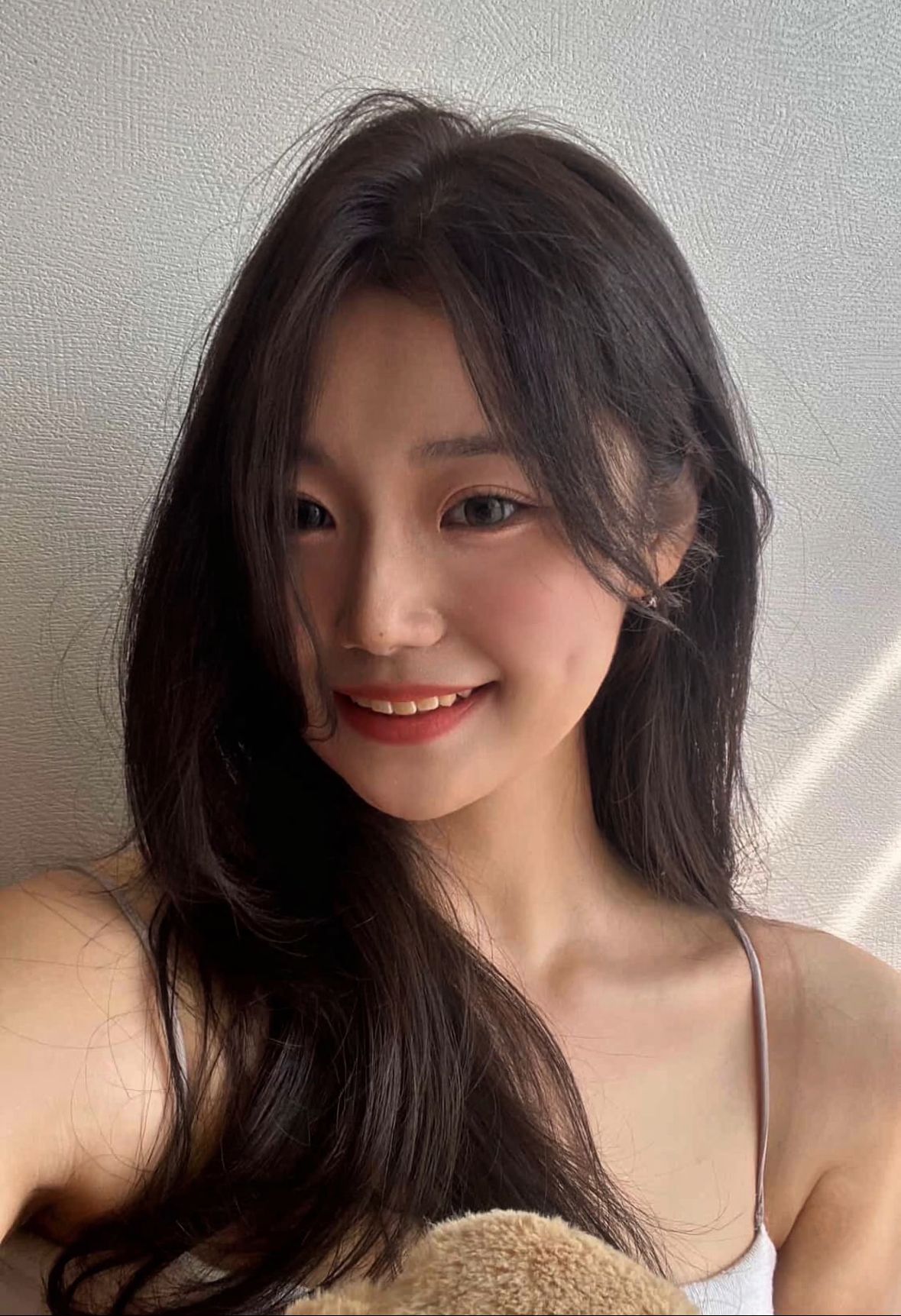 Smiling woman with long dark hair in a light tank top, standing against a textured wall in soft sunlight