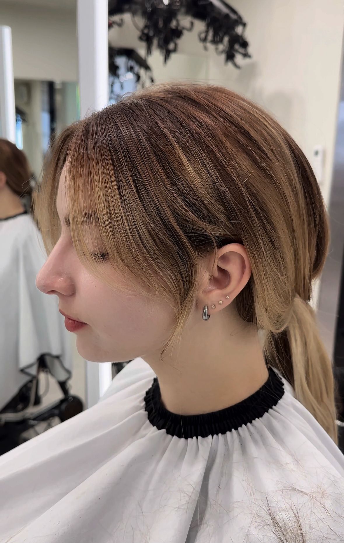Side-profile portrait of a person with layered blonde hair in a salon chair, wearing a white cape.