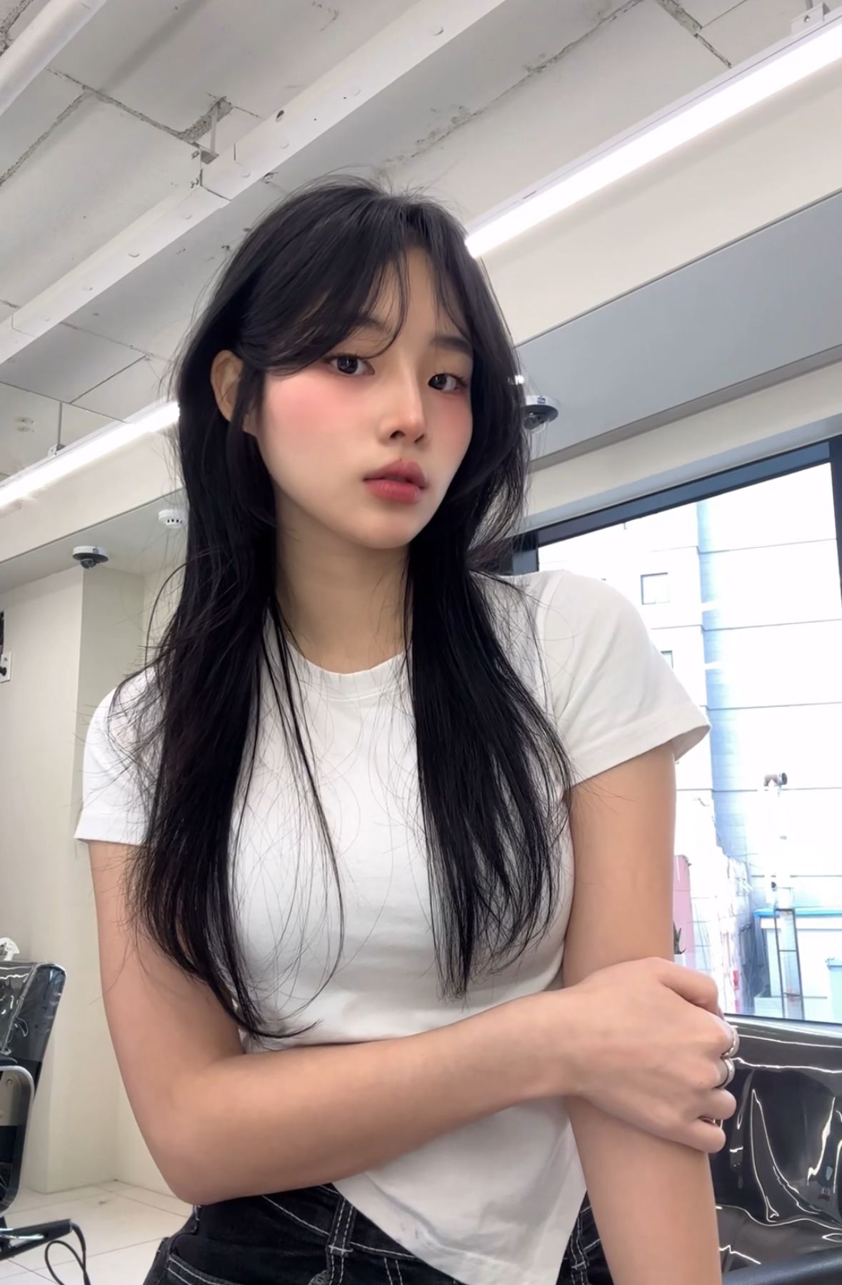 Young woman with long black hair in a white T-shirt standing indoors near windows and a desk
