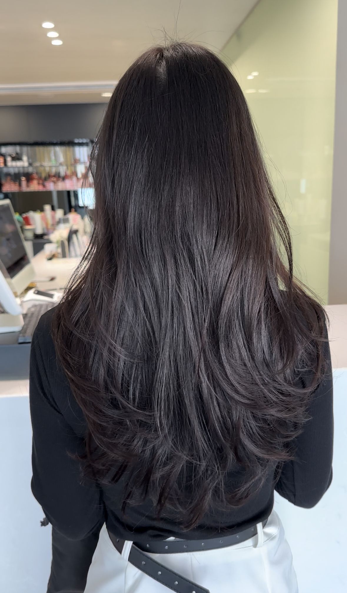 Woman with long dark hair seen from behind in a salon, wearing a black top.