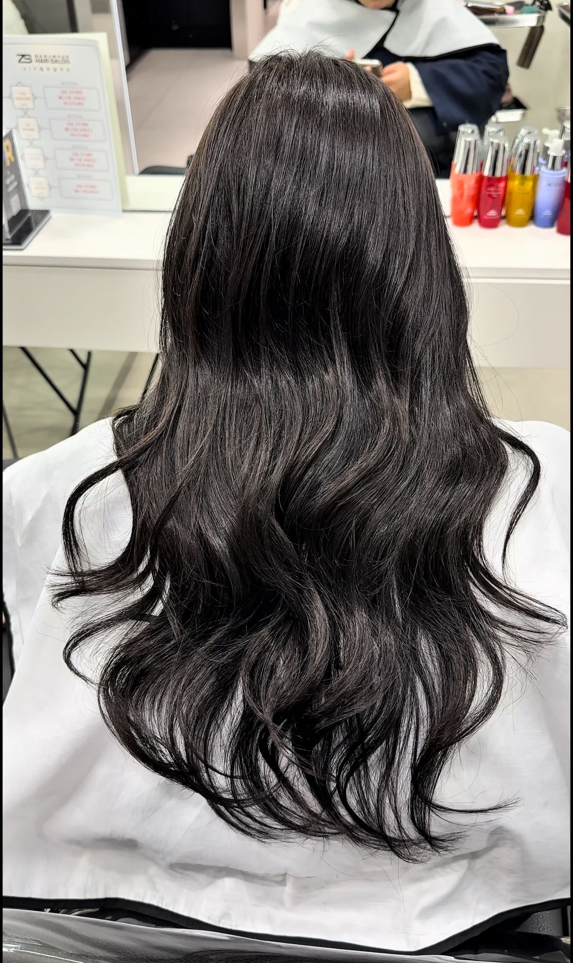 Long, dark wavy hair shown from behind in a salon chair with white cape on