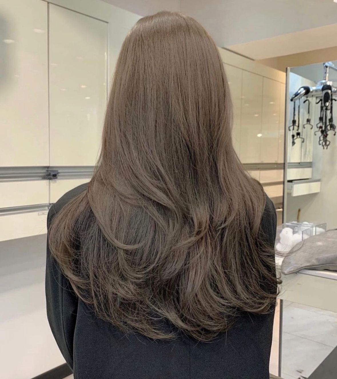 Person with long layered brown hair seen from behind in a salon mirror area