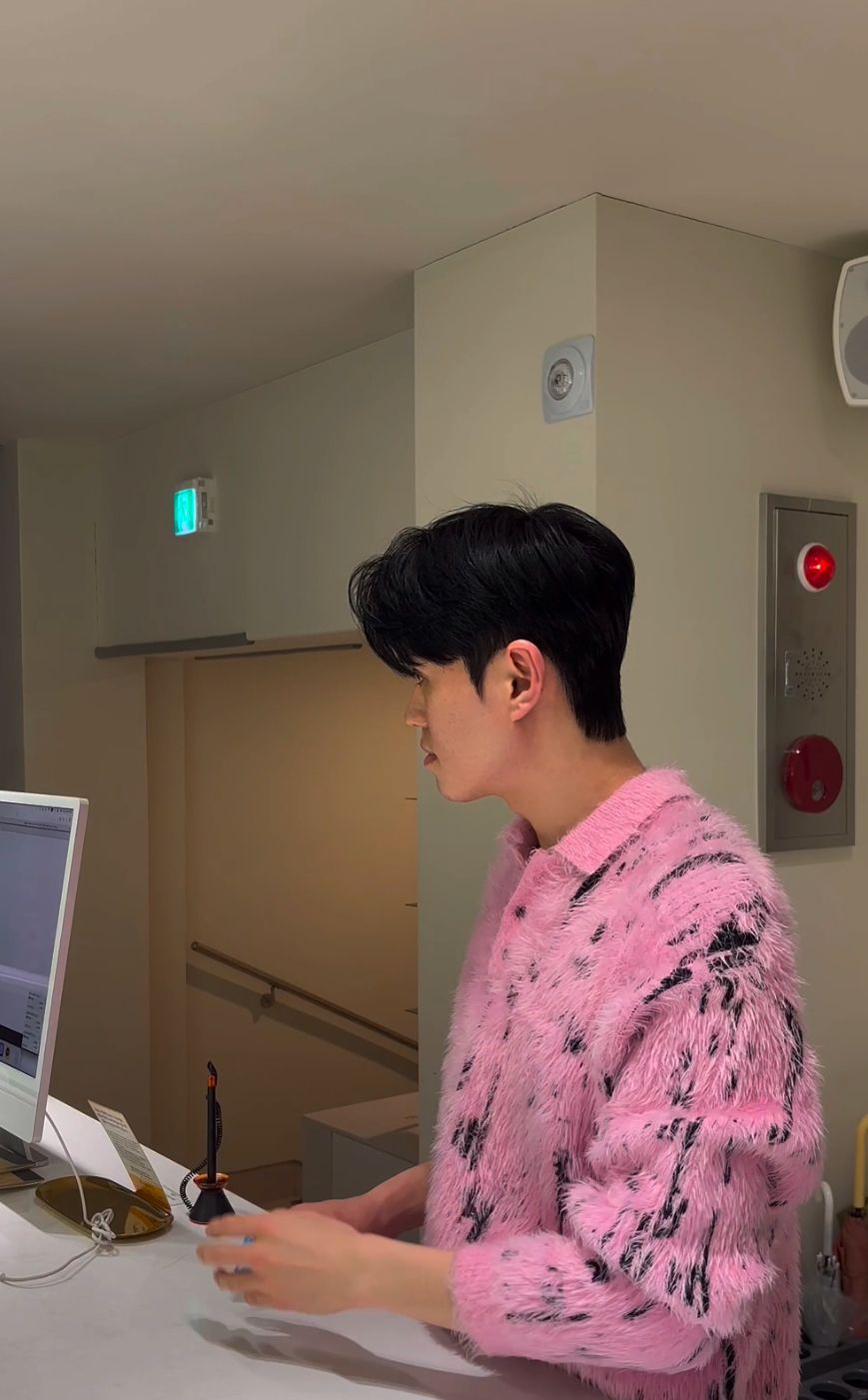 Person in a pink patterned shirt standing at a counter in a hallway, looking to the side.