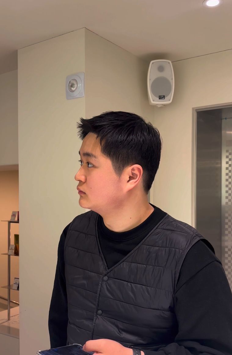 Person in a black quilted vest looking left in a beige indoor room with speakers on the wall.