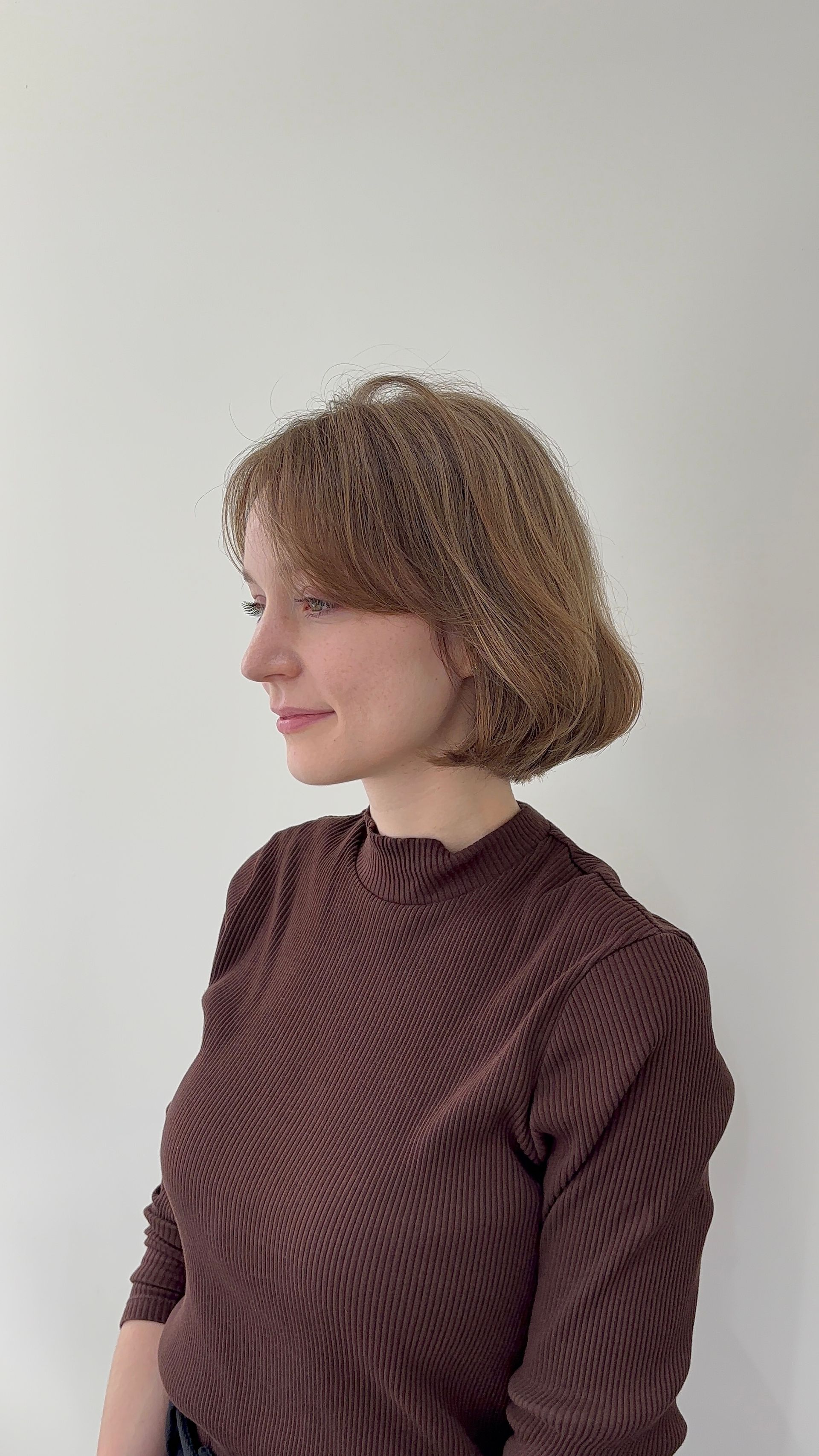 Profile view of a person with short curly hair wearing a brown sweater against a plain wall