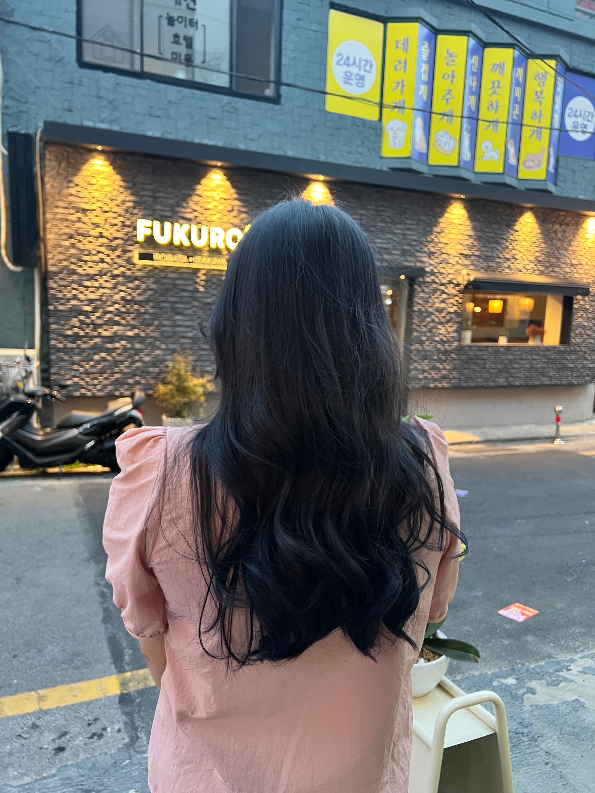 Woman with long dark hair facing a lit storefront at dusk, wearing a pink top and holding a white bag.