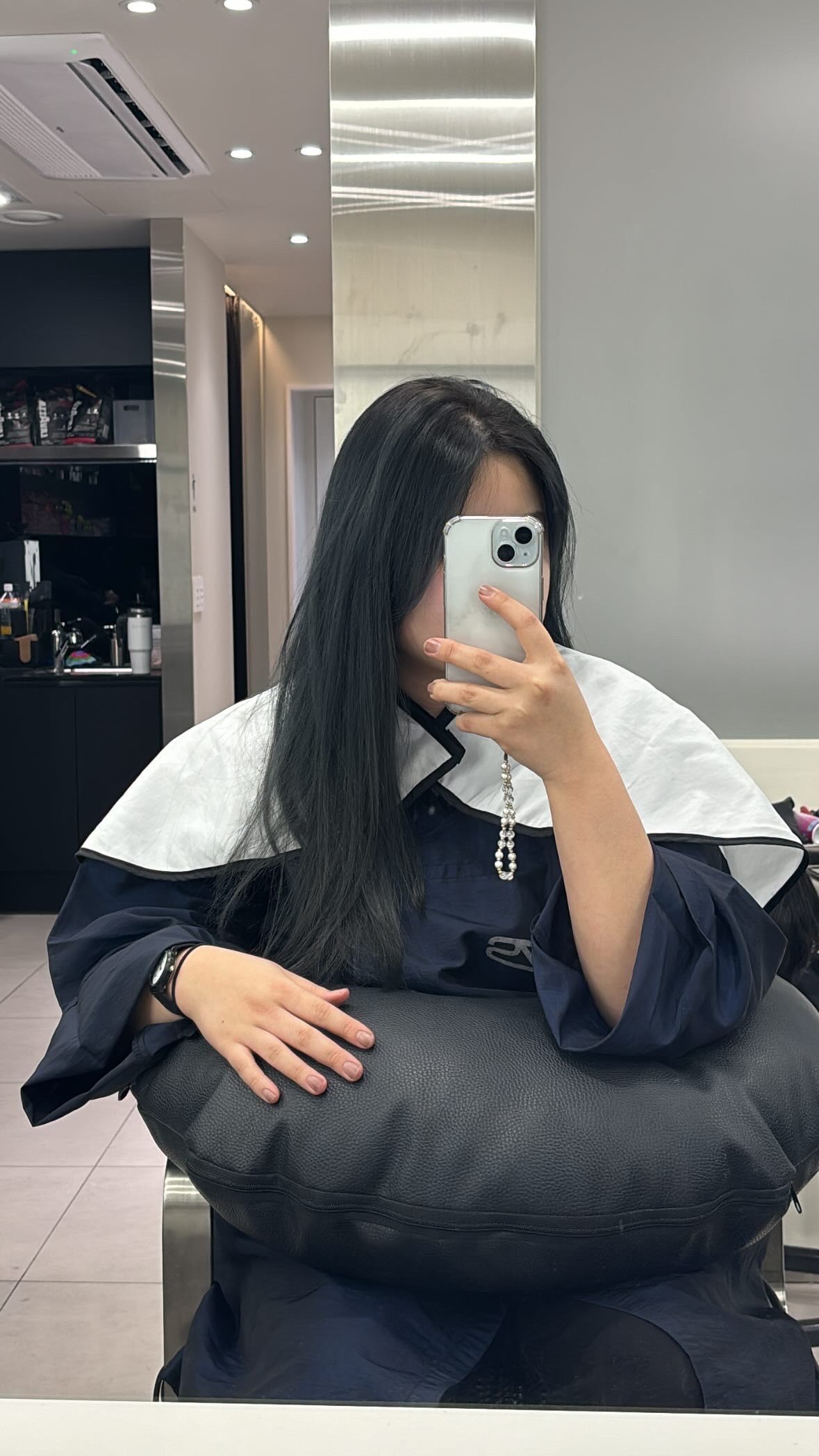 Person with long dark hair taking a mirror selfie in a salon chair, wearing a dark top and white cape.