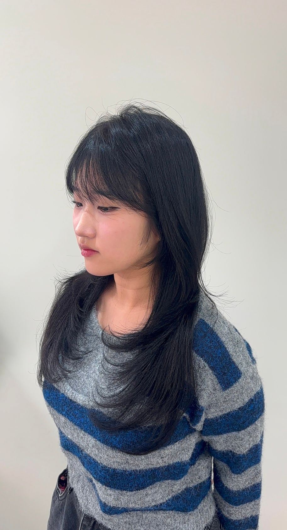 Side profile of a person with long dark hair wearing a blue striped sweater against a plain wall.