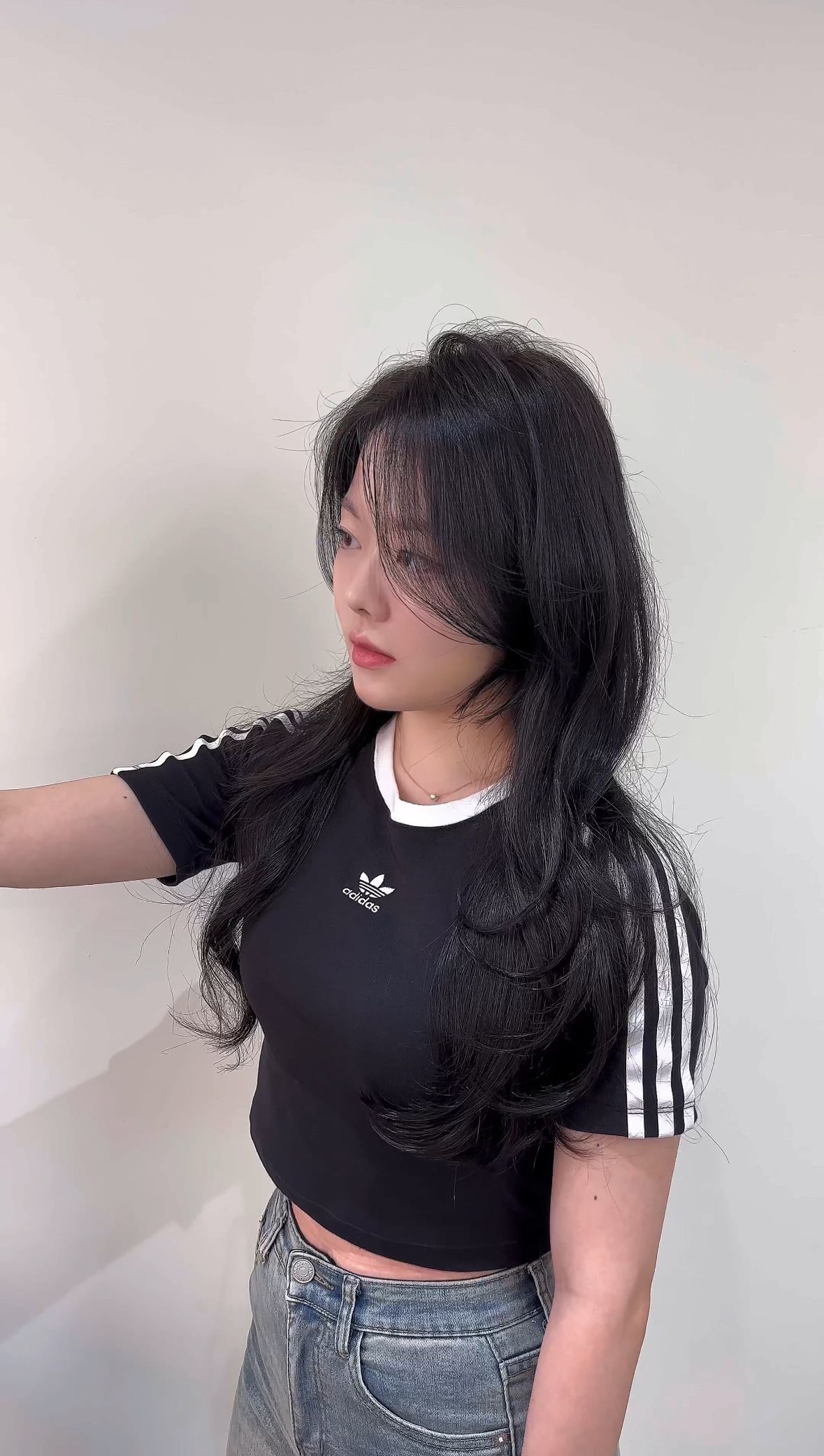 Woman in a black top and jeans posing sideways against a plain wall.