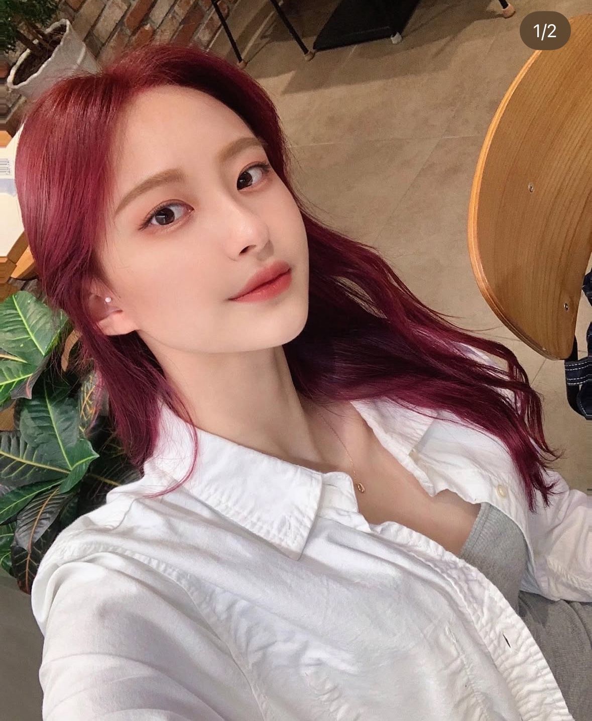 Close-up selfie of a person with burgundy hair, wearing a white shirt, indoors with a wooden chair in the background