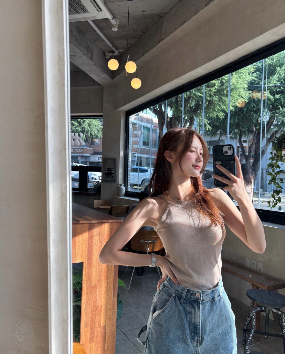 Woman taking a mirror selfie in a bright café, wearing a beige top and blue jeans.