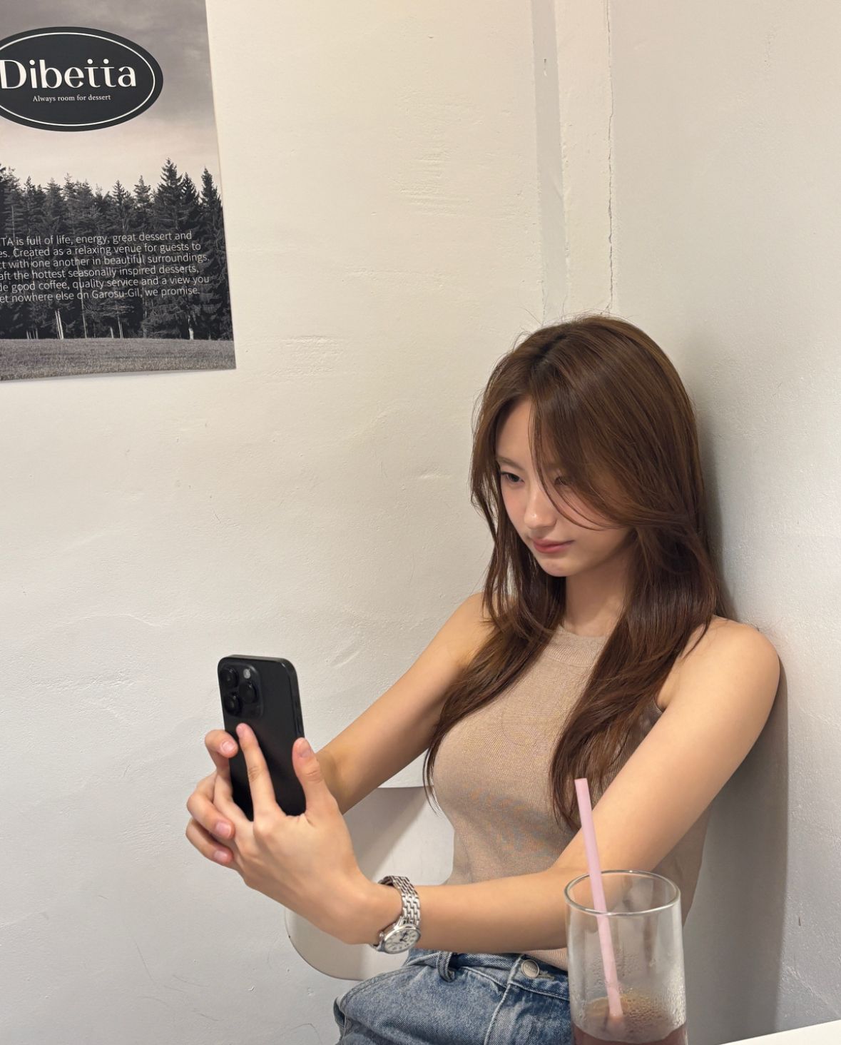 Woman taking a selfie at a café table with a drink and straw, seated by a white wall.