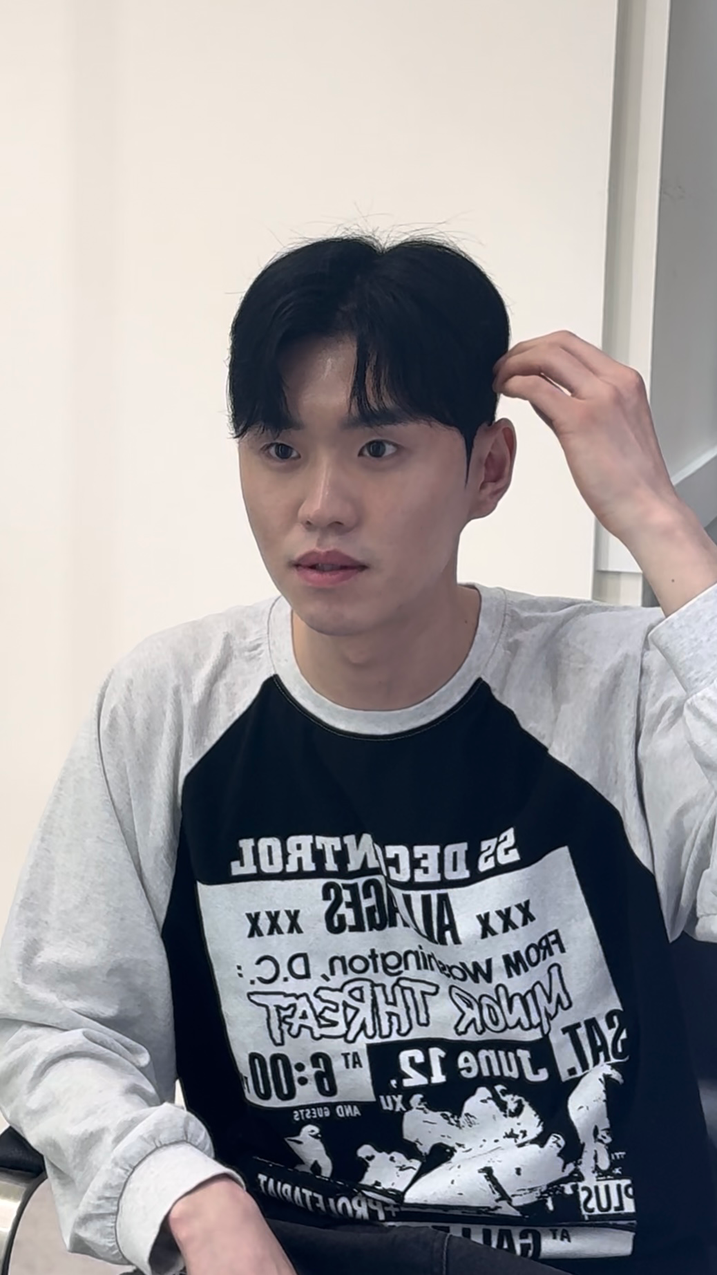 Person in a white long-sleeve shirt with a black graphic, touching their hair and looking aside indoors.