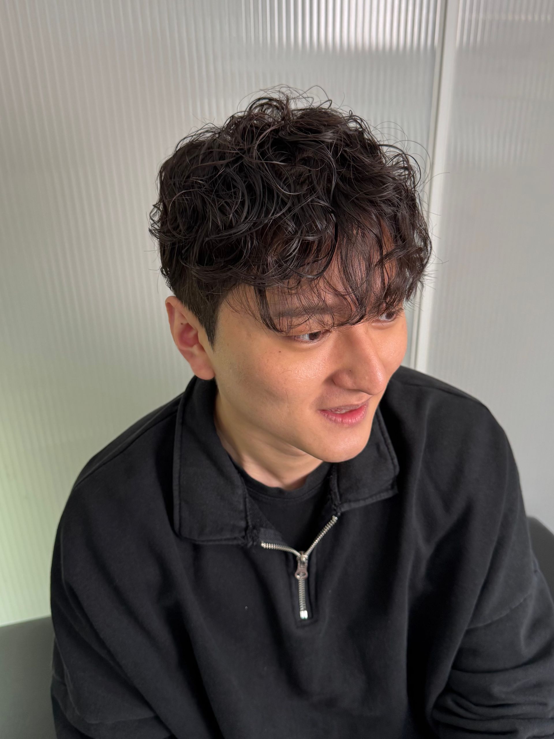 Young person with curly dark hair in a black zip-up jacket, looking down and smiling softly indoors.