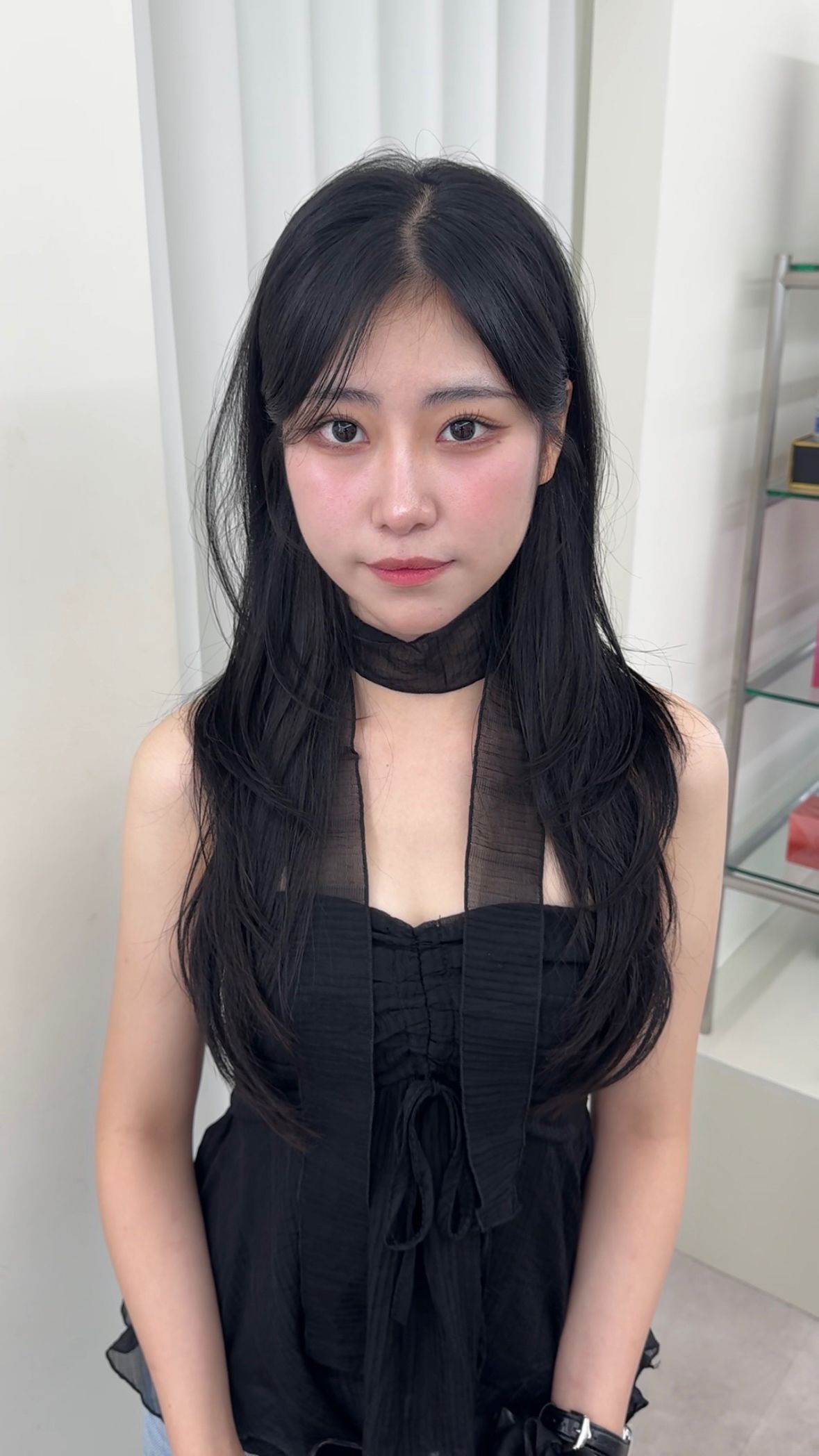 Woman in a black dress with a choker, standing indoors against a light background.