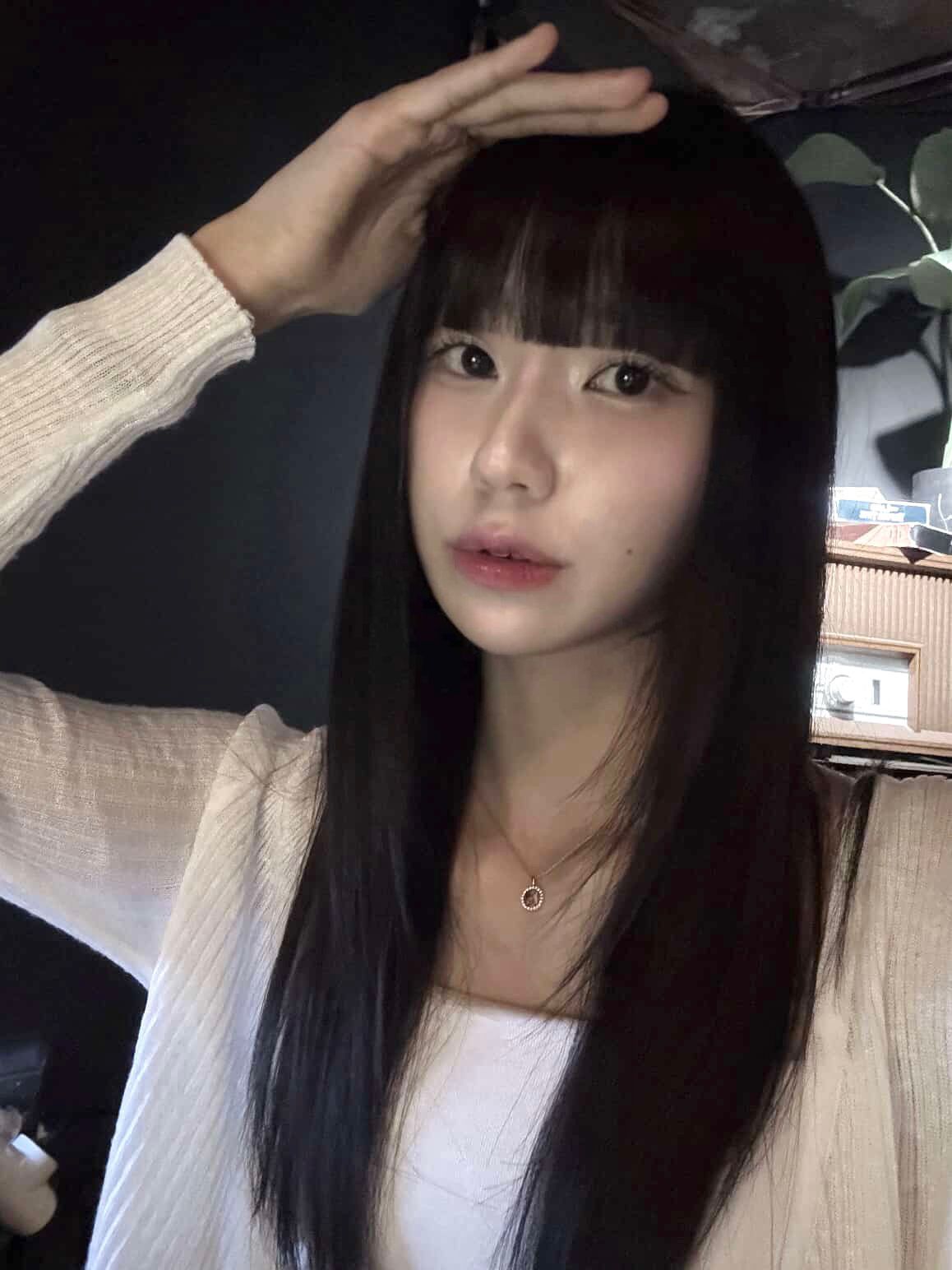 Woman with long dark hair posing indoors, wearing a white top and making a hand salute gesture.