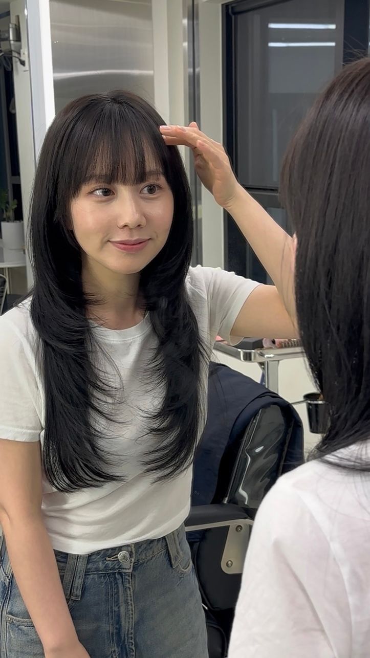 Woman with long black hair adjusting her bangs in a mirror, wearing a white T-shirt and jeans.