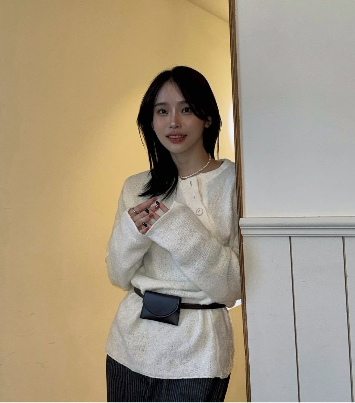 Person in a cream sweater and black belt bag stands by a wall in warm indoor lighting.