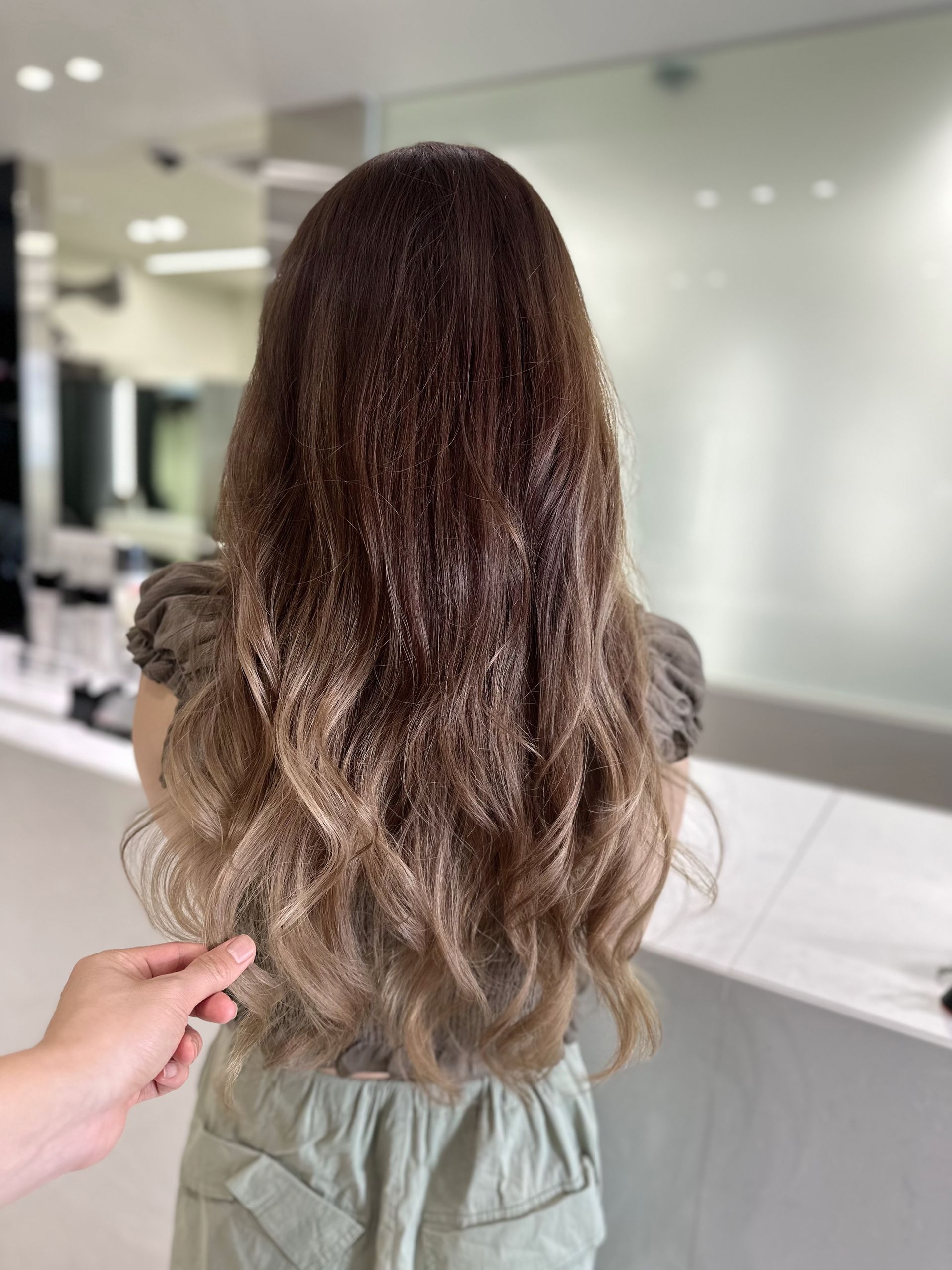 Back view of long wavy brown hair in a salon, with a hand holding the ends to show its length.