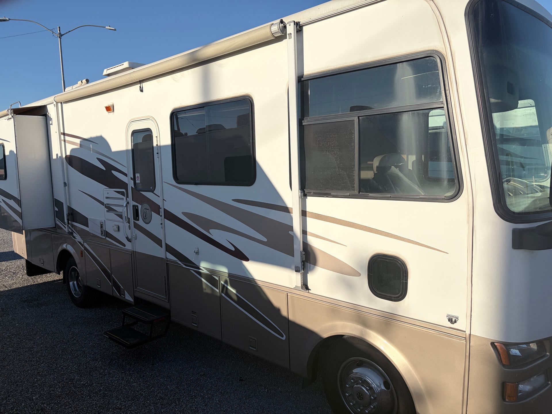 Tan and white RV with slide-out extended, parked on gravel, under a blue sky.
