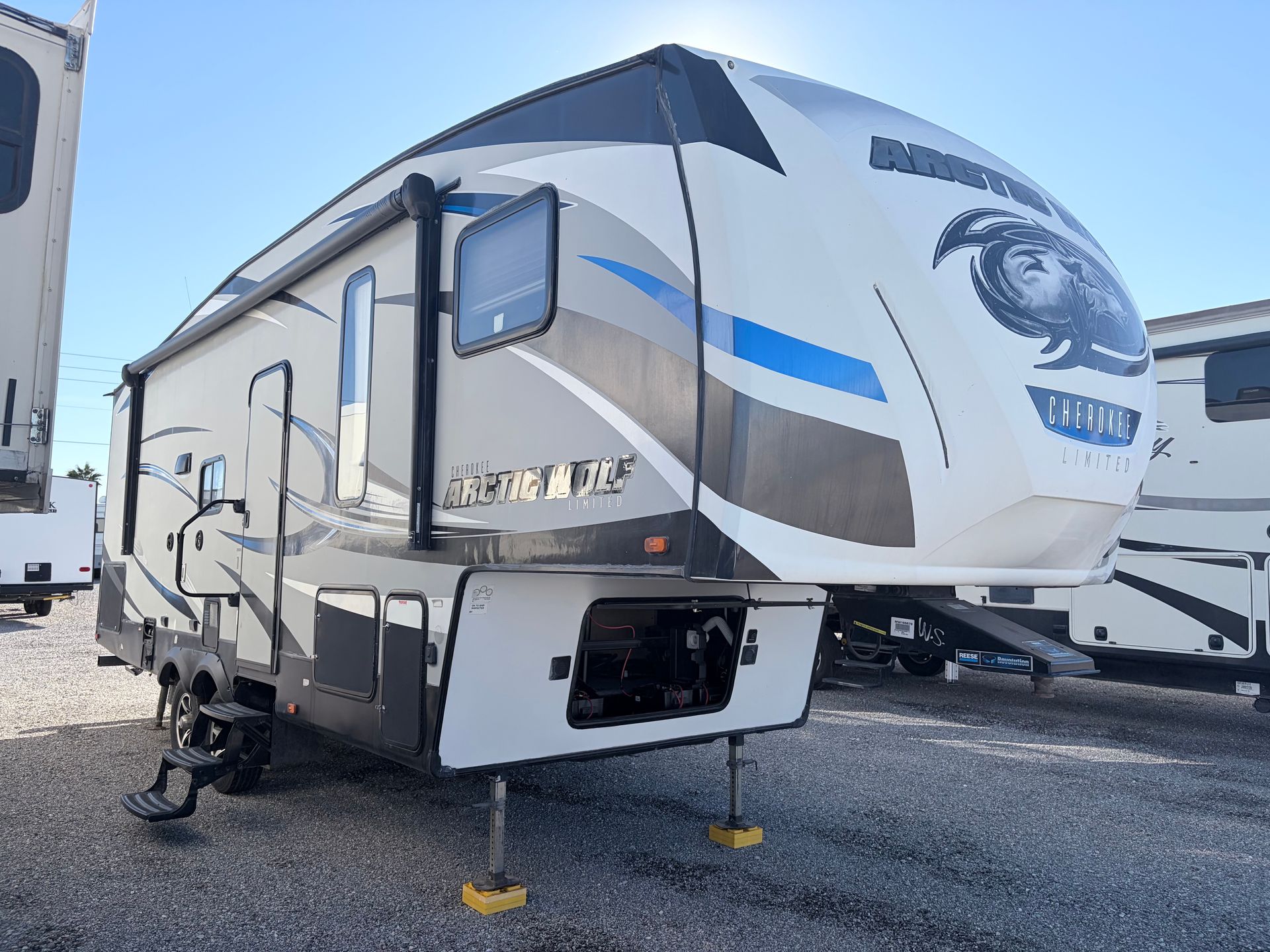A gray and white fifth-wheel RV with blue accents parked on gravel under a bright sky.