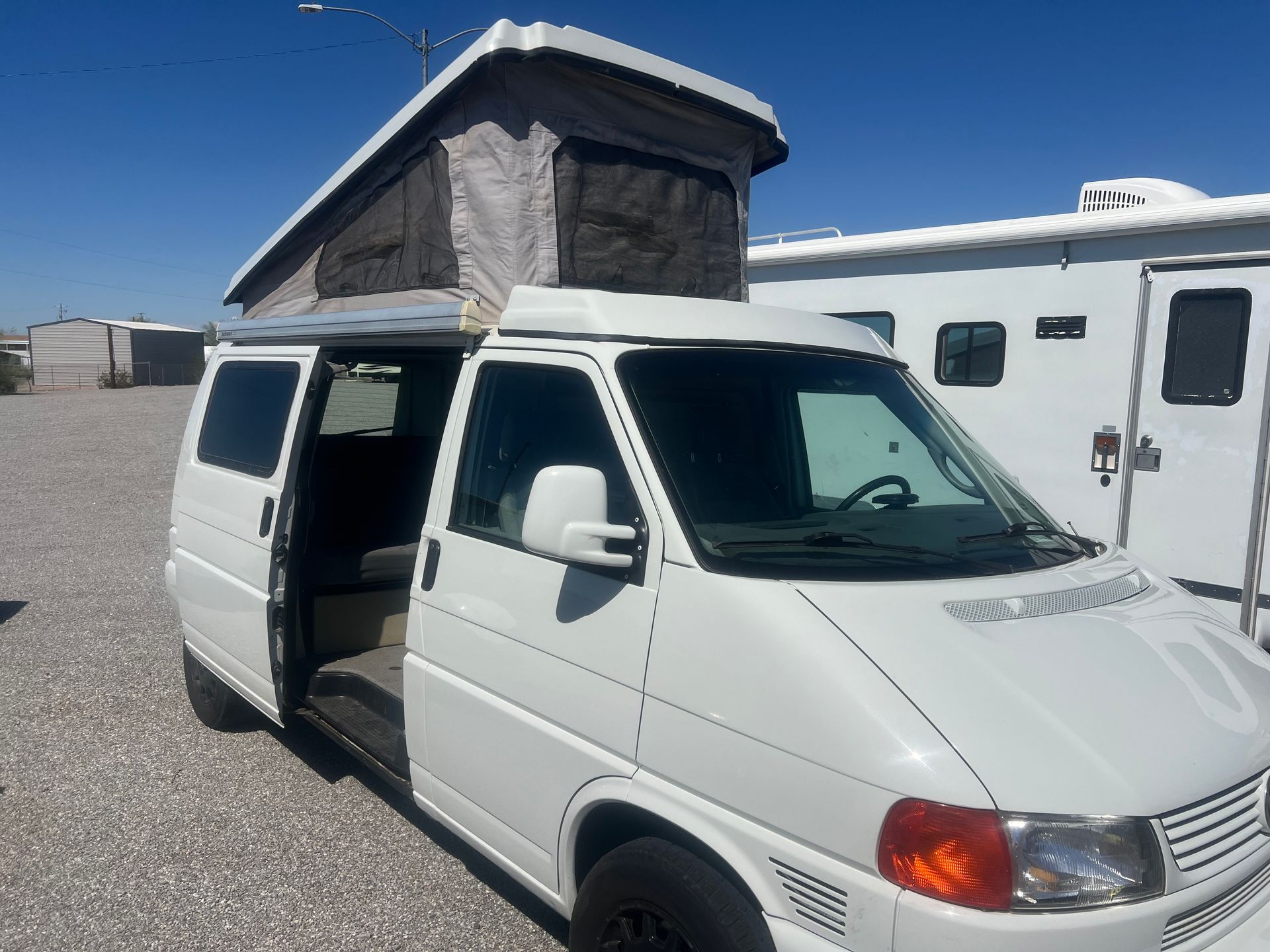 White camper van with pop-up roof, open door. Parked on gravel under a blue sky.