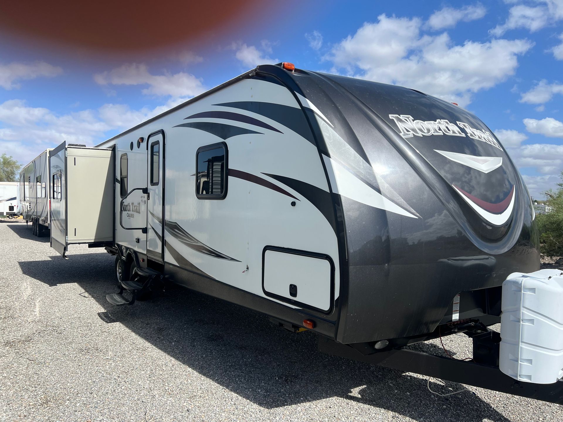 Gray and black Heartland RV trailer with an open awning parked on gravel.