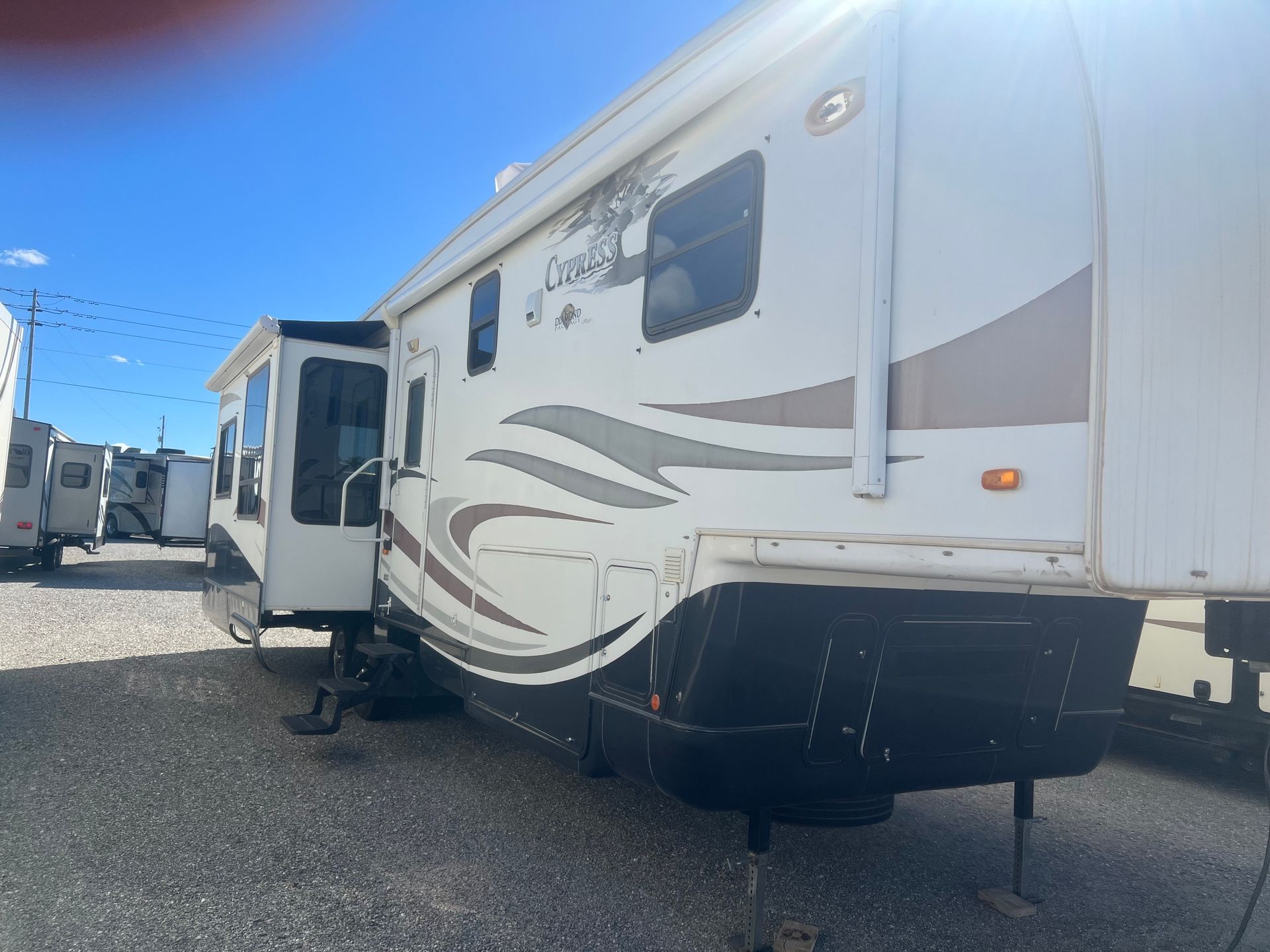 RV trailer, white with brown and gray accents, parked outdoors, slide-out extended, under a bright sky.
