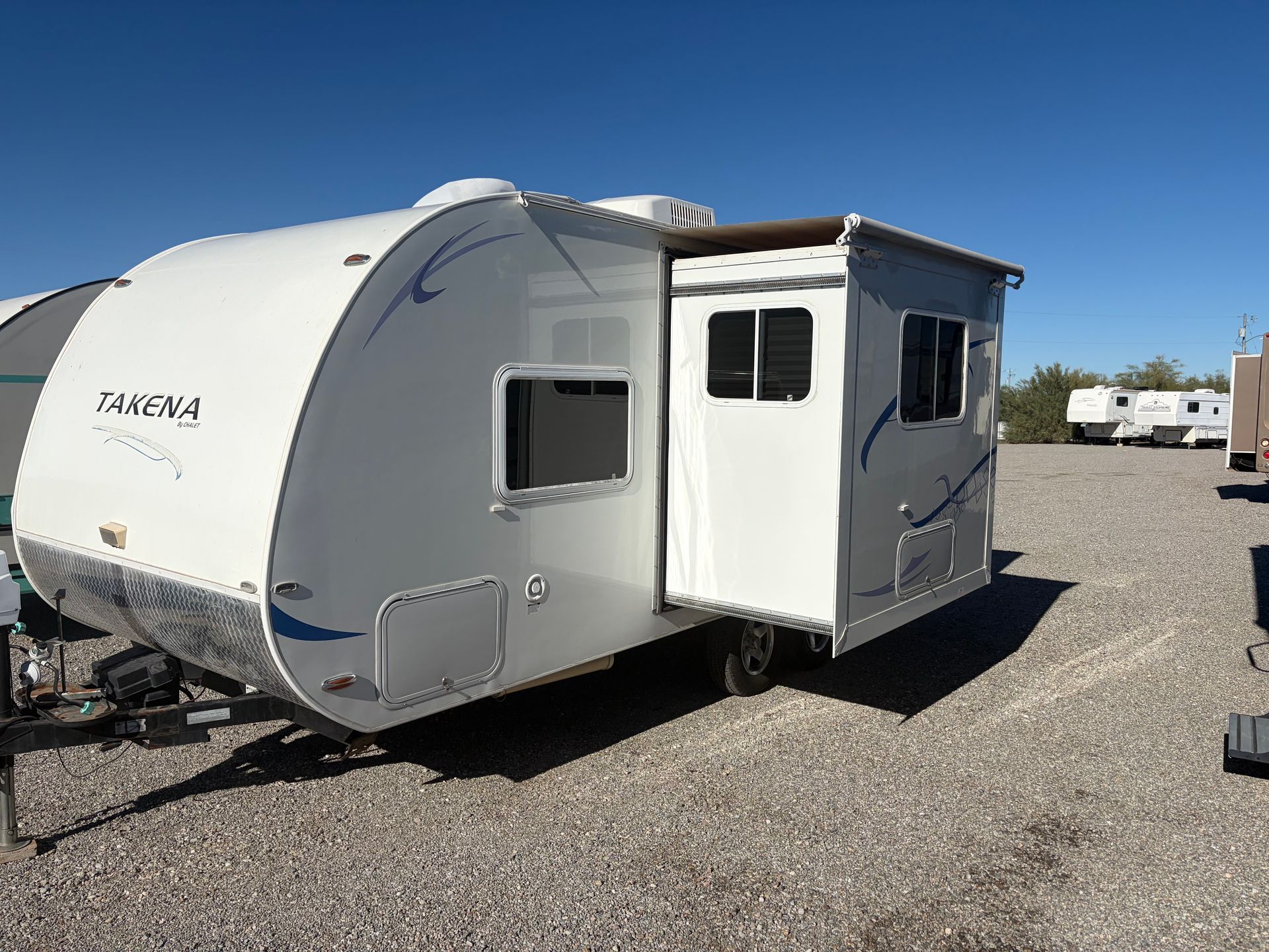 White and blue travel trailer with a slide-out, parked on gravel in a sunny outdoor setting.