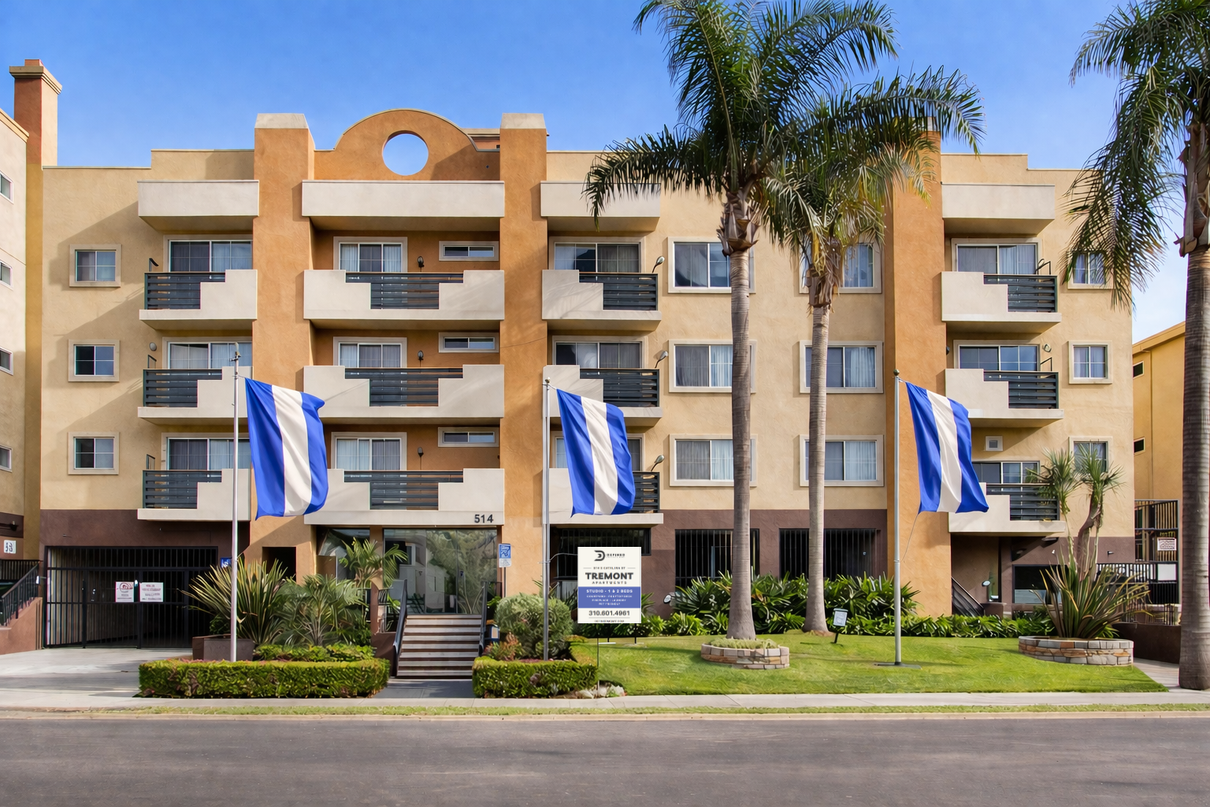 A tan multi-story apartment building with balconies, three vertical blue and white striped flags, and palm trees in front.