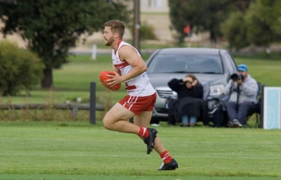 Henry from HL Performance playing AFL, running with a red Sherrin. He wears red and white guernsey, shorts and socks.