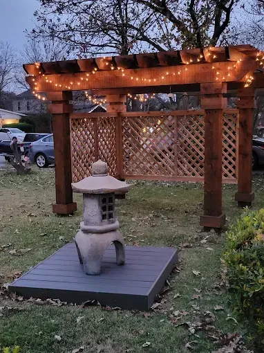 A Japanese garden scene with a stone lantern, wooden pergola with string lights, and lattice.