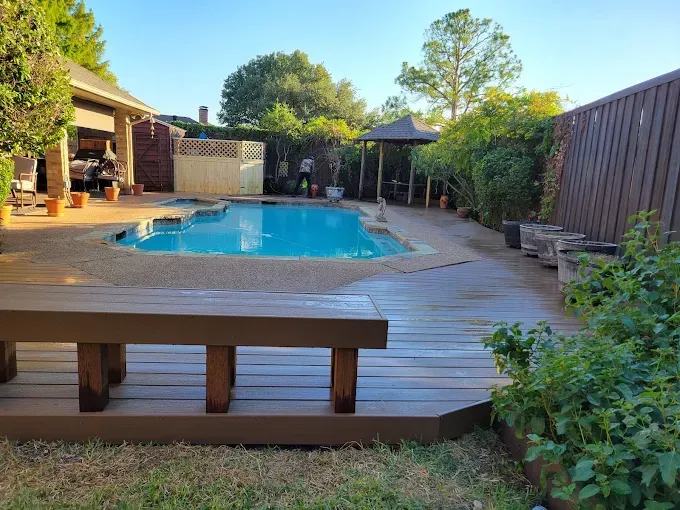 Backyard pool with wooden deck and bench, surrounded by greenery and a gazebo.