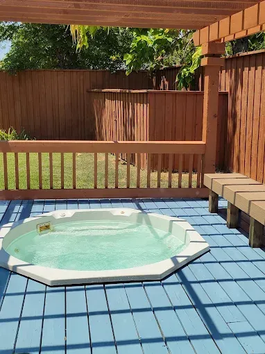 Hot tub on a blue deck, with a wooden pergola, benches, and a brown fence in the background.