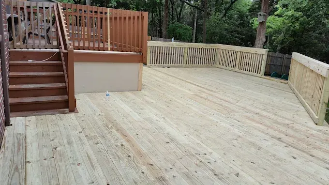 A wooden deck with stairs, light-colored wood, and a brown railing. Green trees in the background.