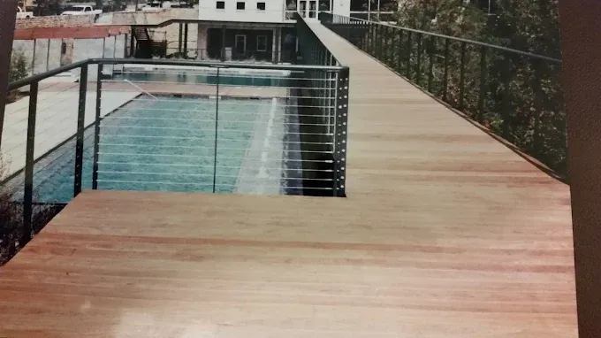 Wooden deck with stainless steel railing overlooks a rectangular pool with a building in the background.