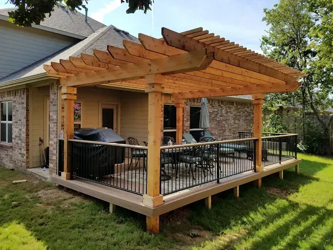 Wooden pergola with black railing on a deck, extending from a house, surrounded by grass.