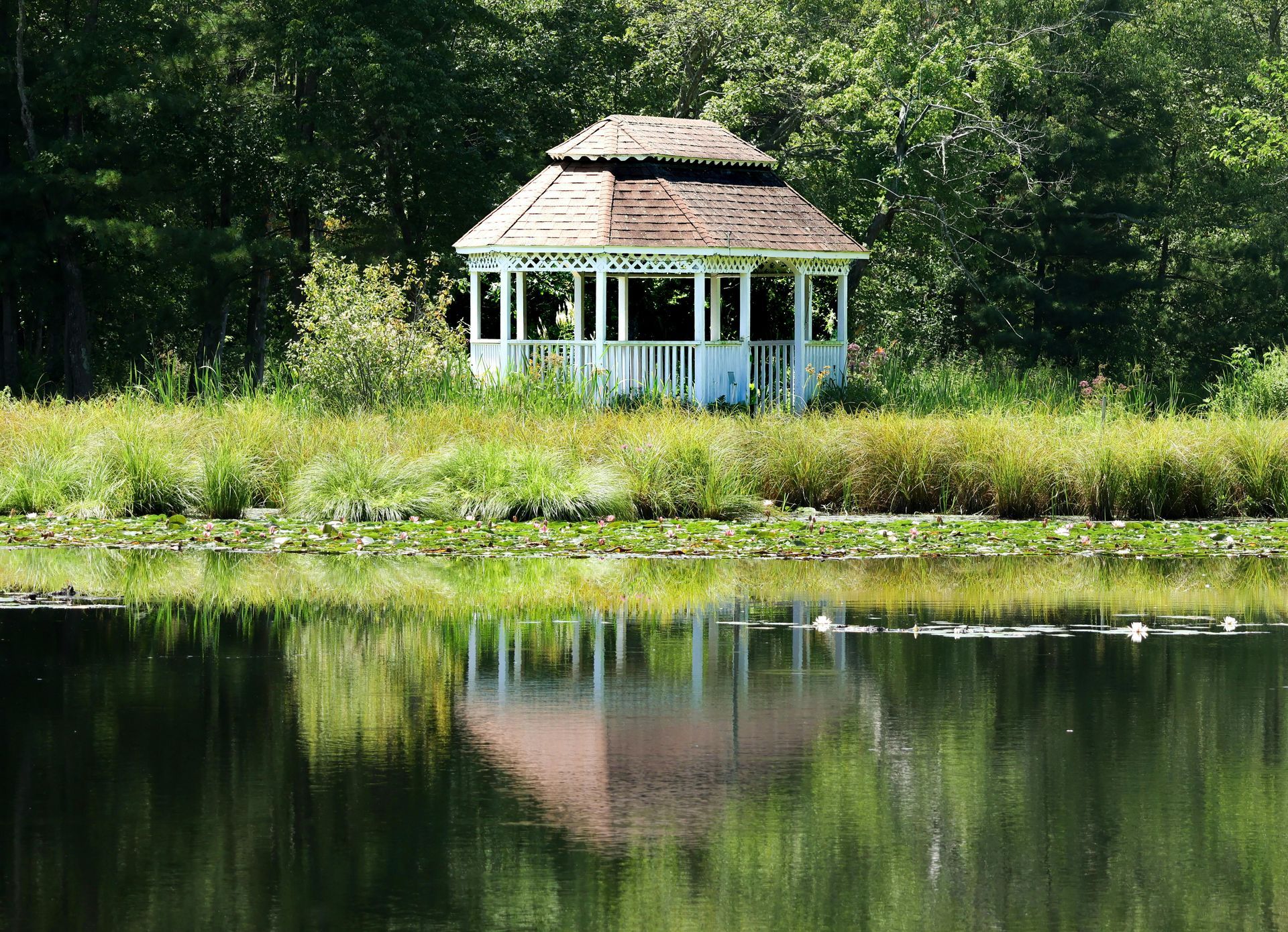White gazebo on a grassy bank, reflecting in a calm lake surrounded by green foliage.