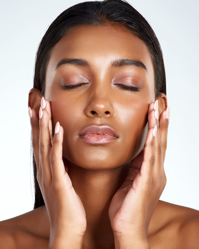 Woman with closed eyes, gently touching cheeks with palms. Skin appears dewy, against a light backdrop.