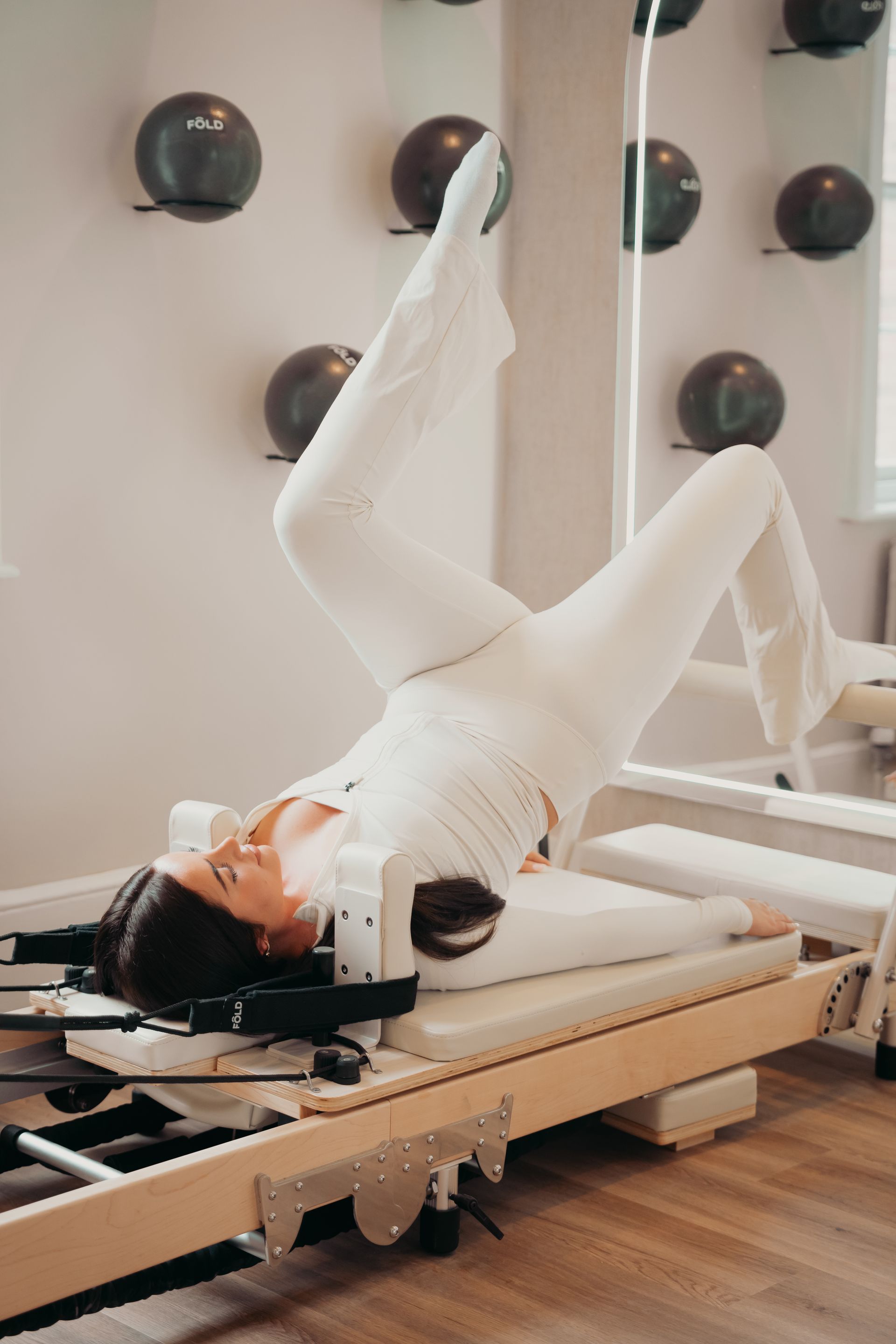 Woman doing Pilates on a reformer, legs in straps, arms raised. Neutral setting, beige tones.