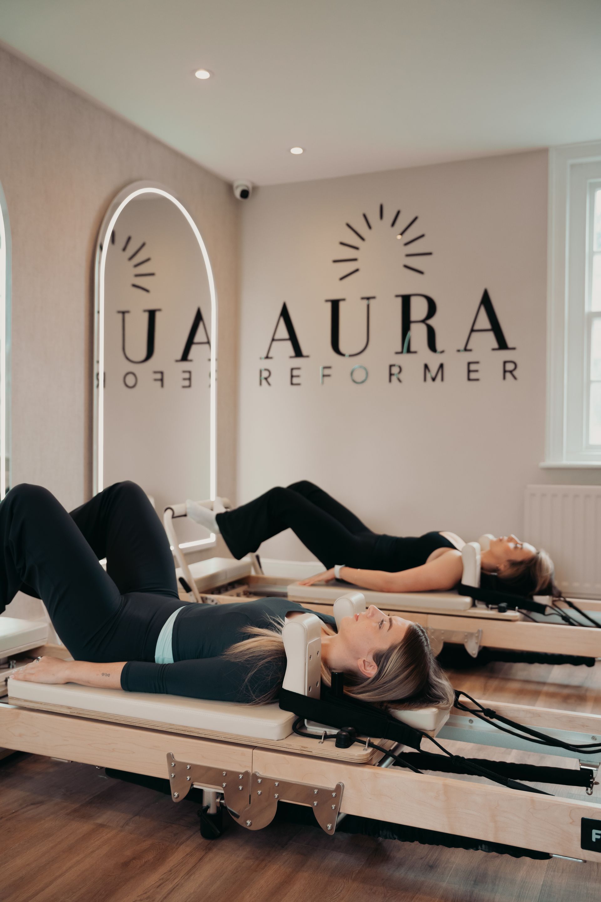 Woman doing Pilates on a reformer, legs in straps, arms raised. Neutral setting, beige tones.