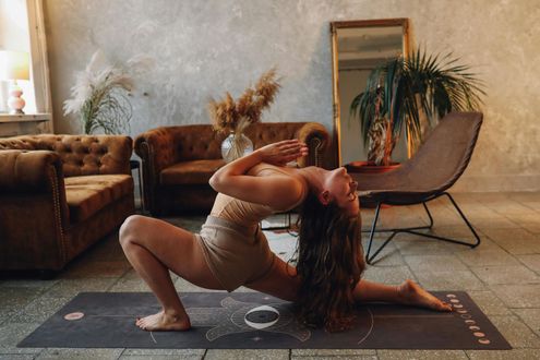 Woman in yoga pose on mat, arms raised, head back, in a living room with brown sofa and chair.