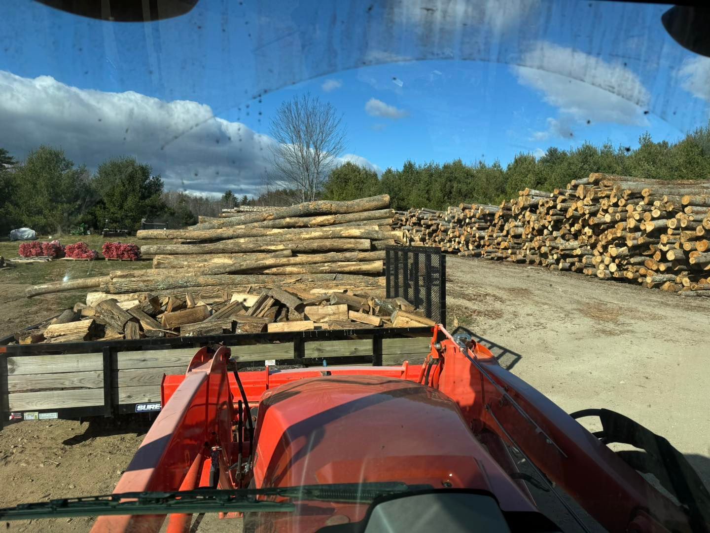 A tractor is driving down a dirt road next to a pile of logs.