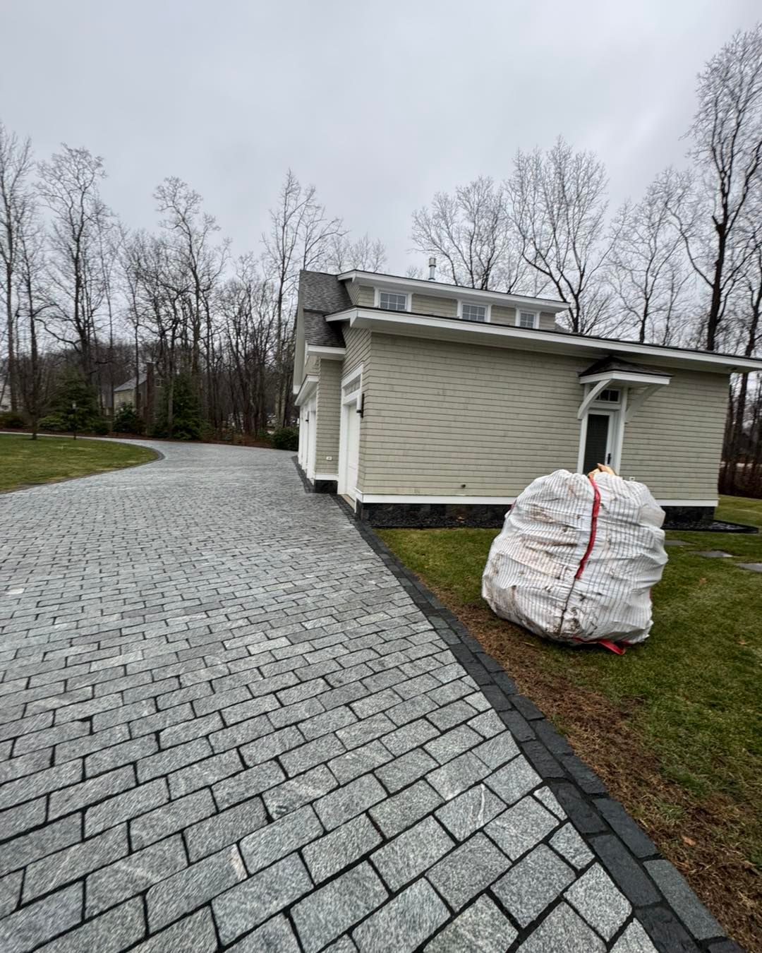 A large bag of trash is sitting on the side of a driveway next to a house.